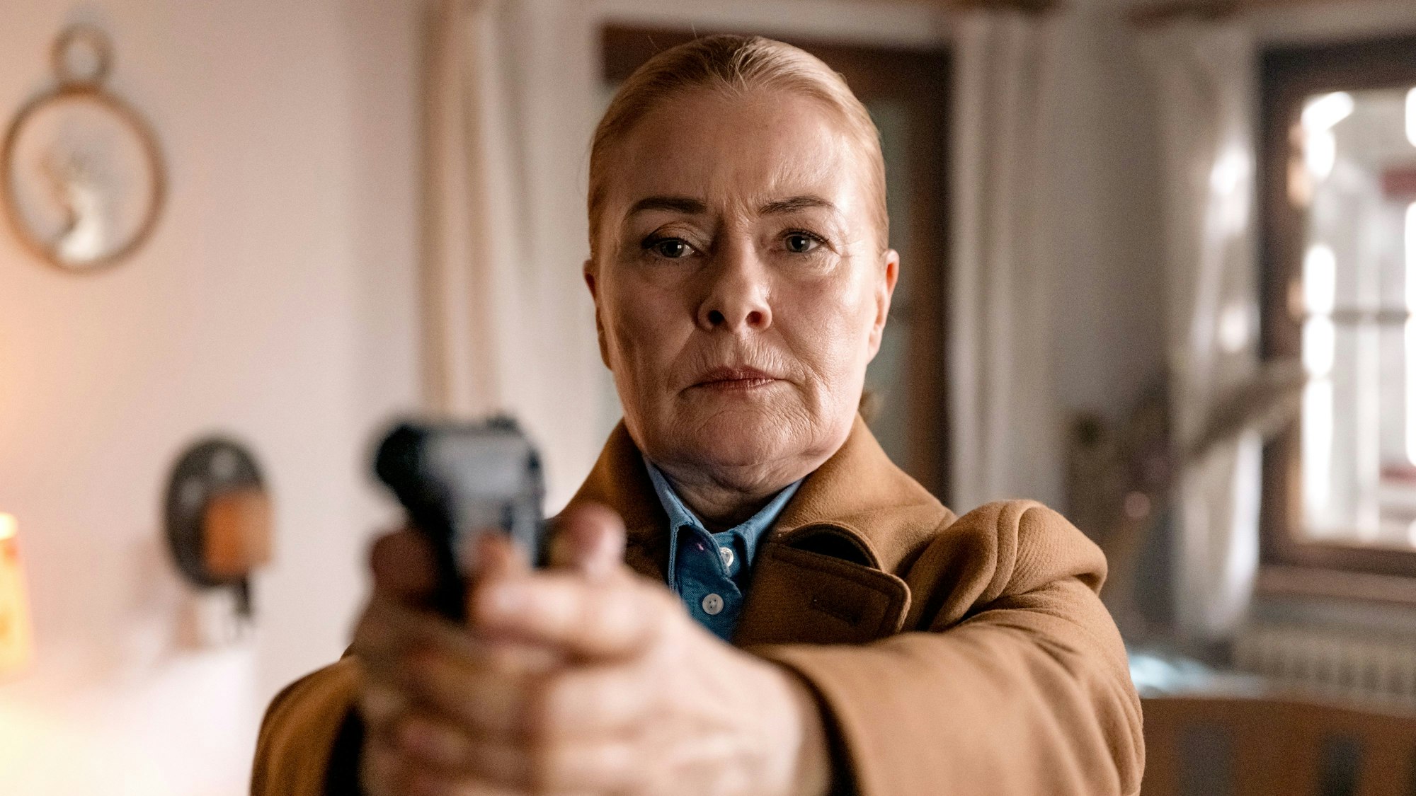 Eine Frau mittleren Alters mit streng zurückgekämmten Haaren richtet eine Pistole in Richtung Kamera.