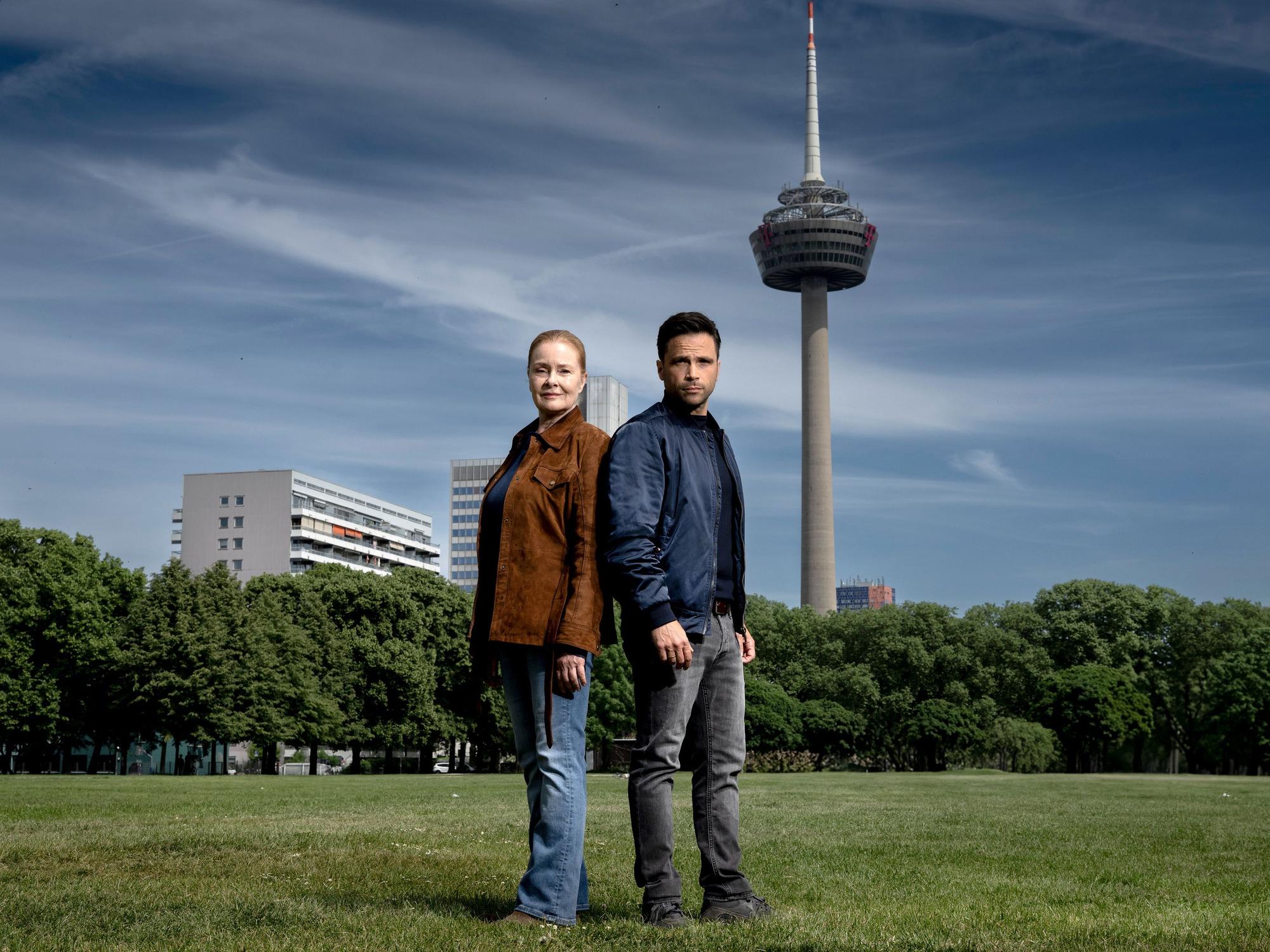 Ein Mann und eine Frau stehen auf einer Wiese, im Hintergrund ragt ein großer Fernsehturm in den blauen Himmel.