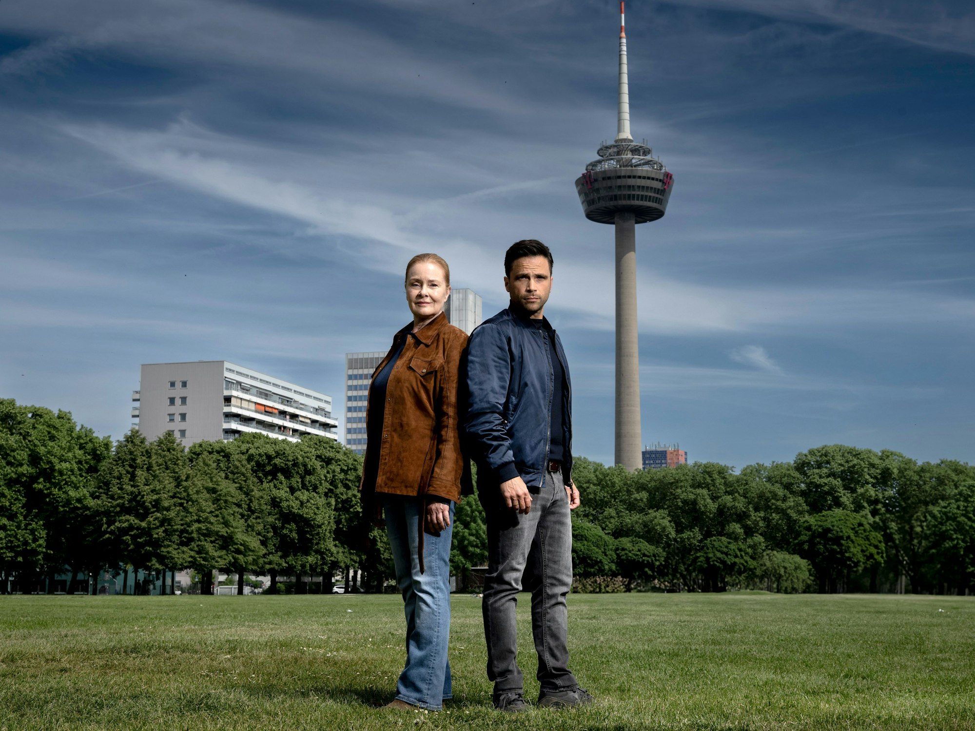 Ein Mann und eine Frau stehen auf einer Wiese, im Hintergrund ragt ein großer Fernsehturm in den blauen Himmel.