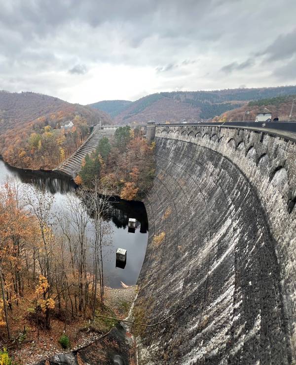 Eine hohe, verwitterte Staumauer steht in einem See, dahinter herbstlich gefärbter Laubwald.