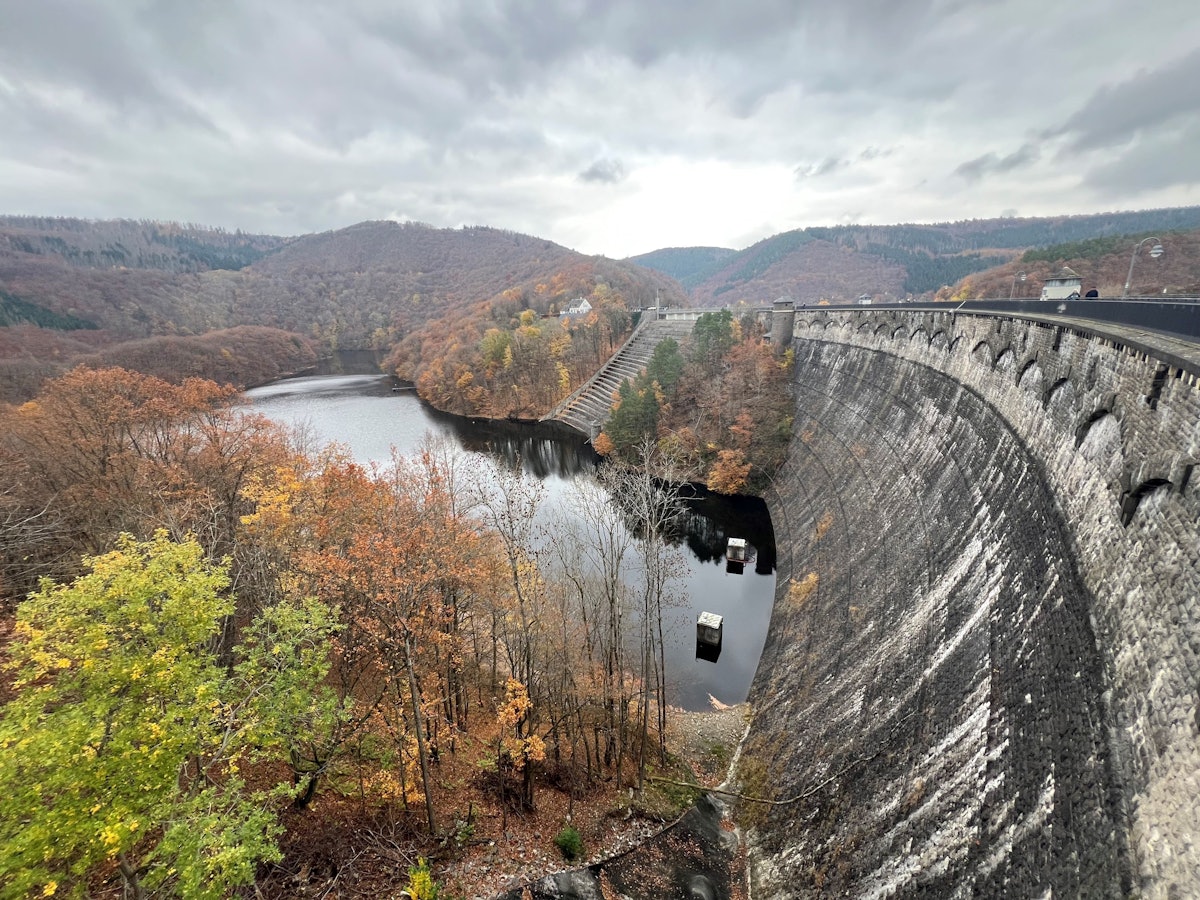 Eine hohe, verwitterte Staumauer steht in einem See, dahinter herbstlich gefärbter Laubwald.