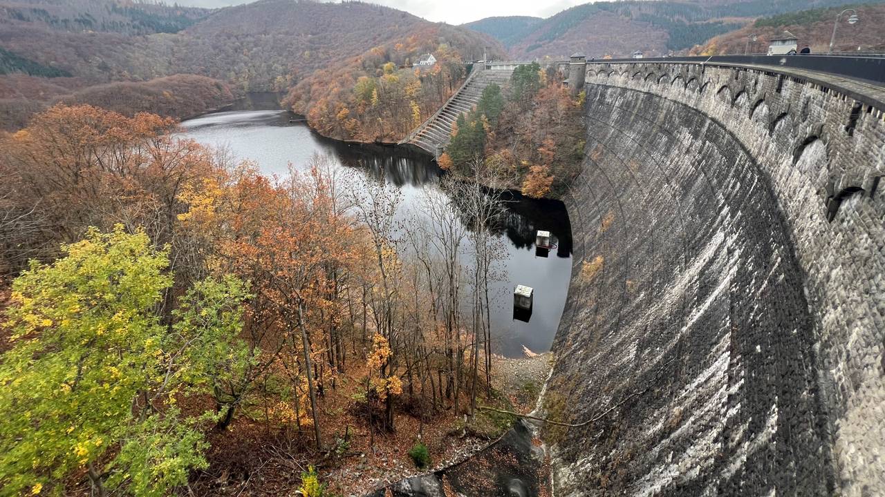 Eine hohe, verwitterte Staumauer steht in einem See, dahinter herbstlich gefärbter Laubwald.