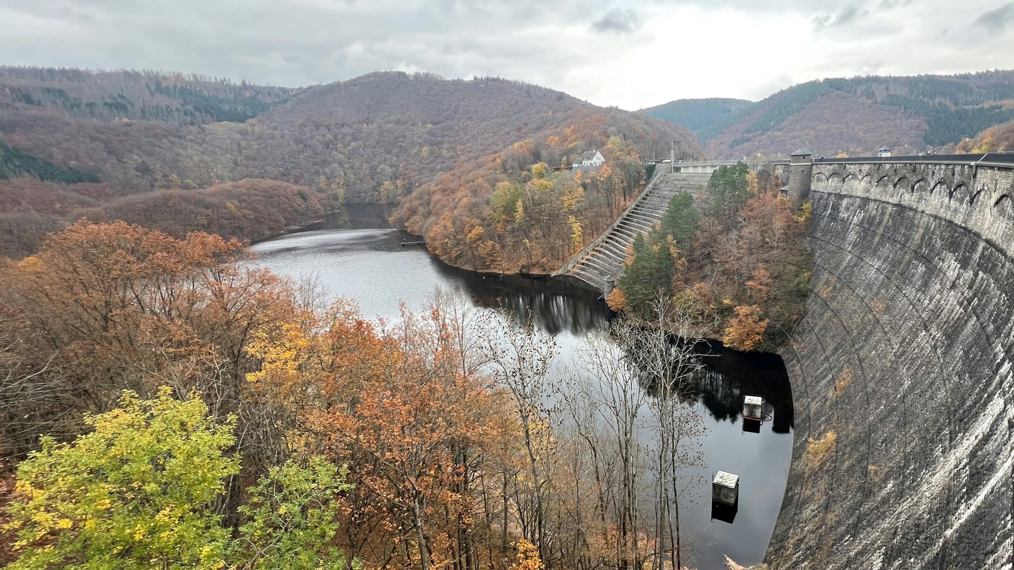 Eine hohe, verwitterte Staumauer steht in einem See, dahinter herbstlich gefärbter Laubwald.
