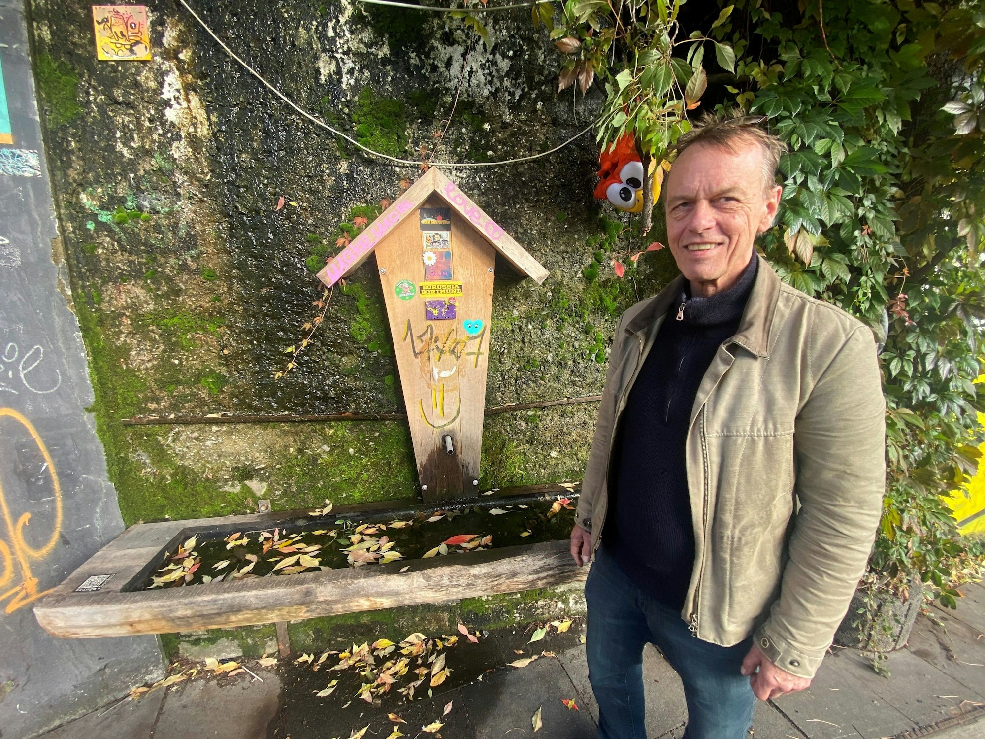 Schreiner Michael Krings steht vor einem alpin gestalteten Brunnen an der einer Brückenmauer