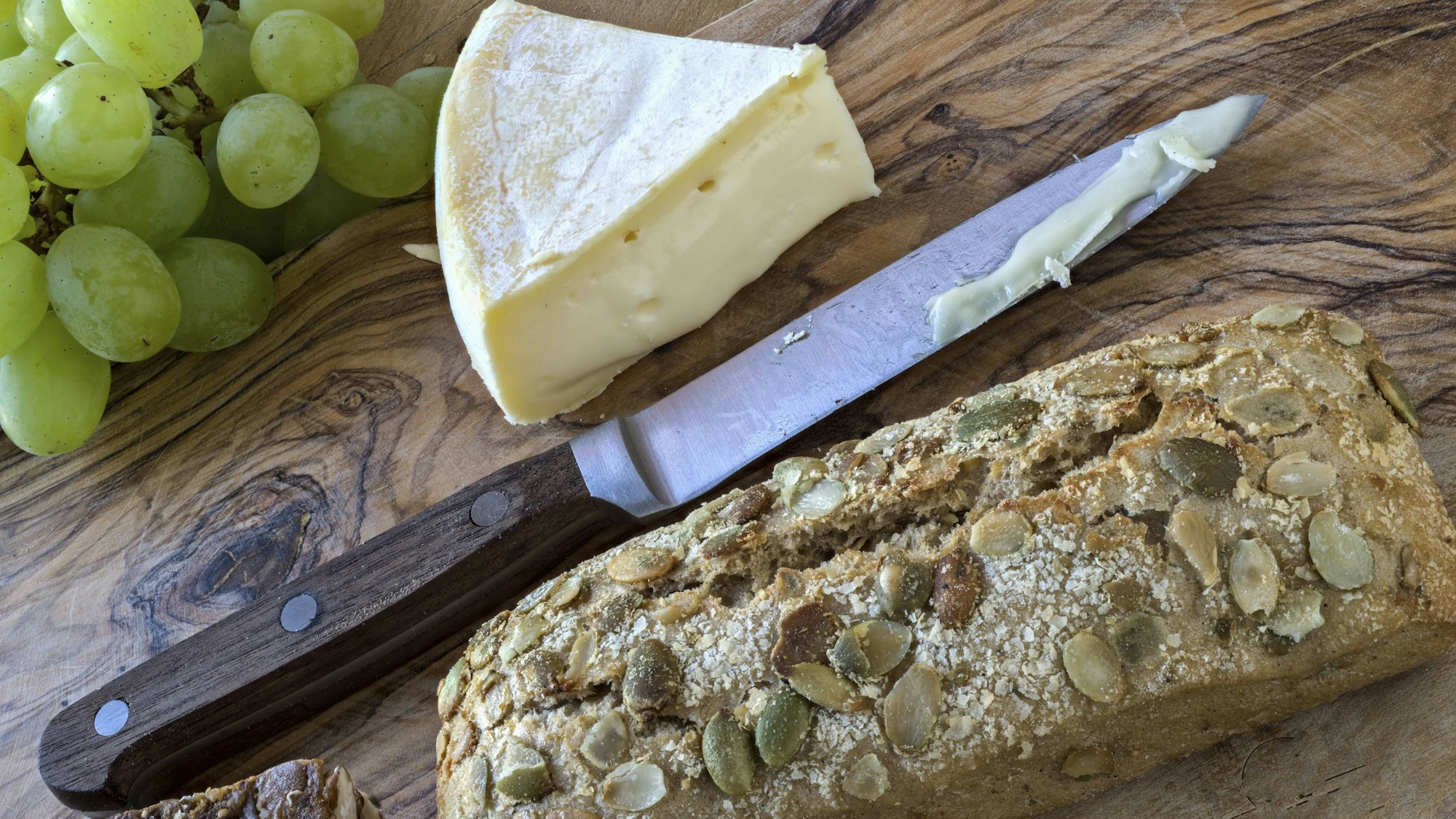 Grüne Trauben, Weichkäse, ein Messer und Körnerbrot auf einem Holzschneidebrett