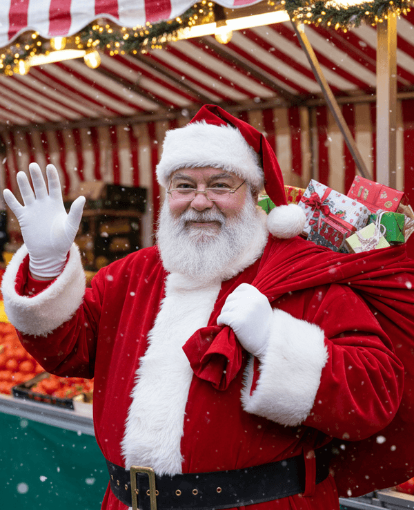 Ein Weihnachtsmann steht vor einem Wochenmarktstand, schaut freundlich in die Kamera und winkt. Auf dem Rücken hat er einen Sack mit kleinen Geschenken.