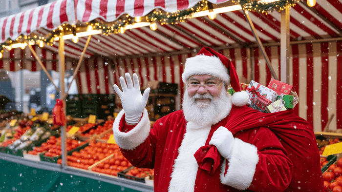 Ein Weihnachtsmann steht vor einem Wochenmarktstand, schaut freundlich in die Kamera und winkt. Auf dem Rücken hat er einen Sack mit kleinen Geschenken.