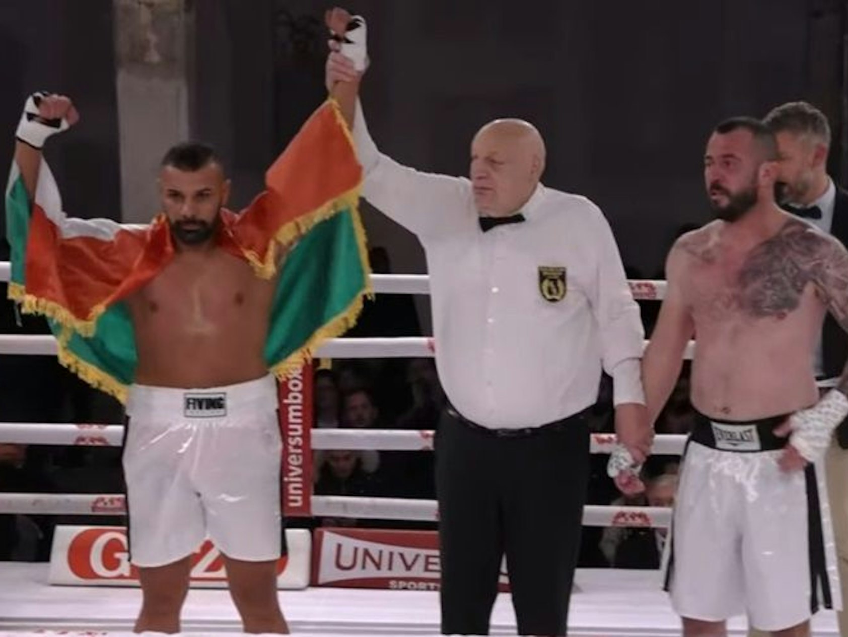 Ein Mann steht zwischen zwei Boxern und hält dem einen den Arm als Zeichen des Sieges hoch.