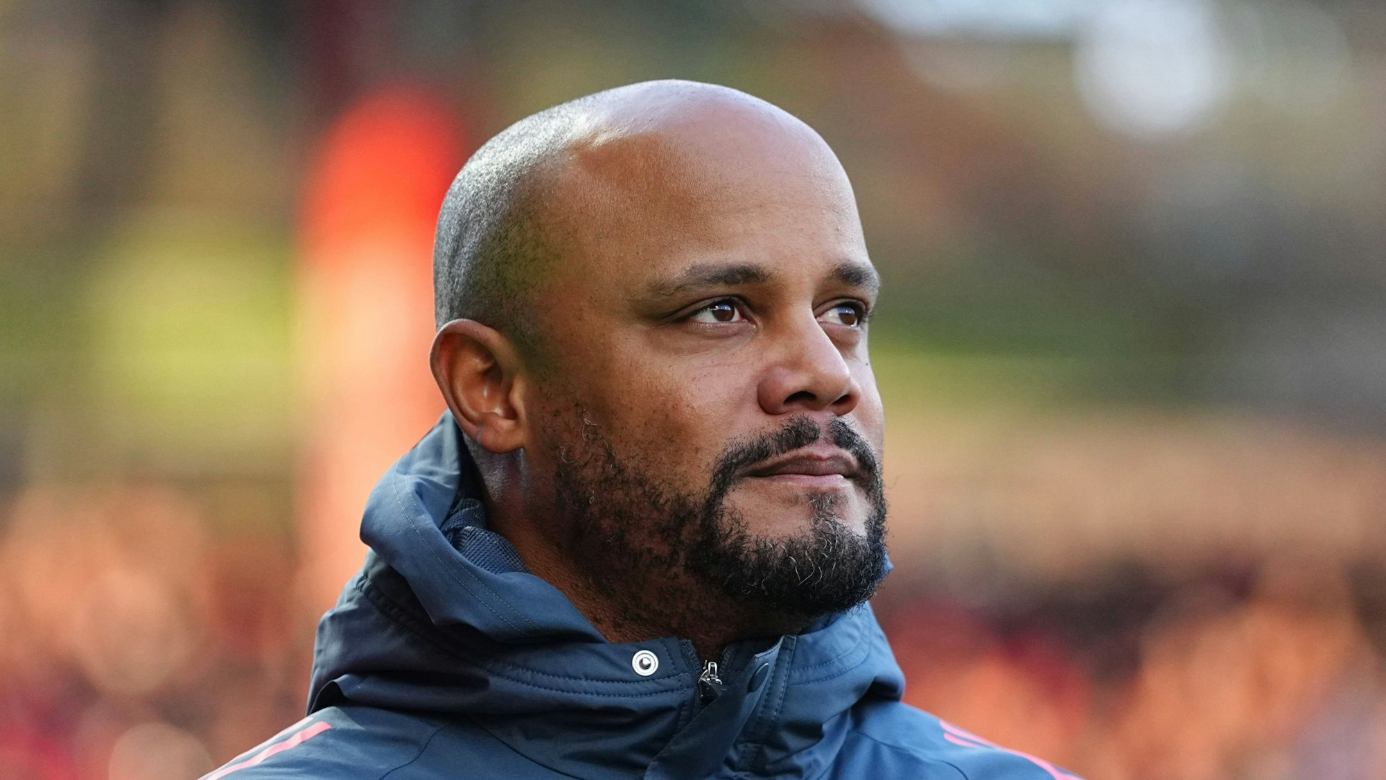 Vincent Kompany mit Bart und rasierter Glatze, blickt im Profil in blauer Jacke