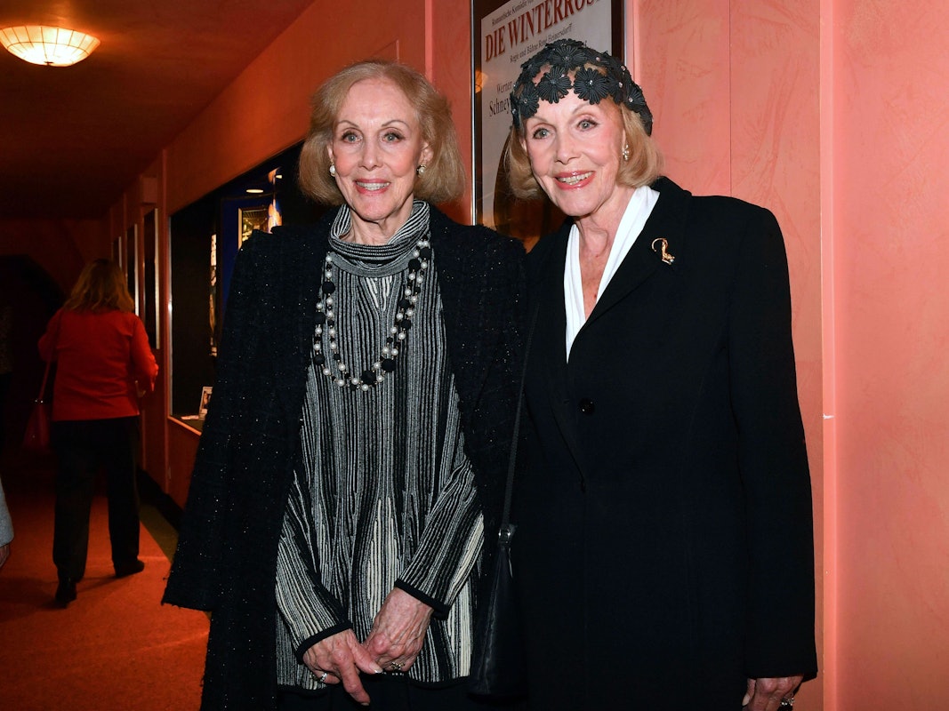 Zwei lächelnde ältere Frauen stehen nebeneinander vor einem Plakat in einem Innenraum