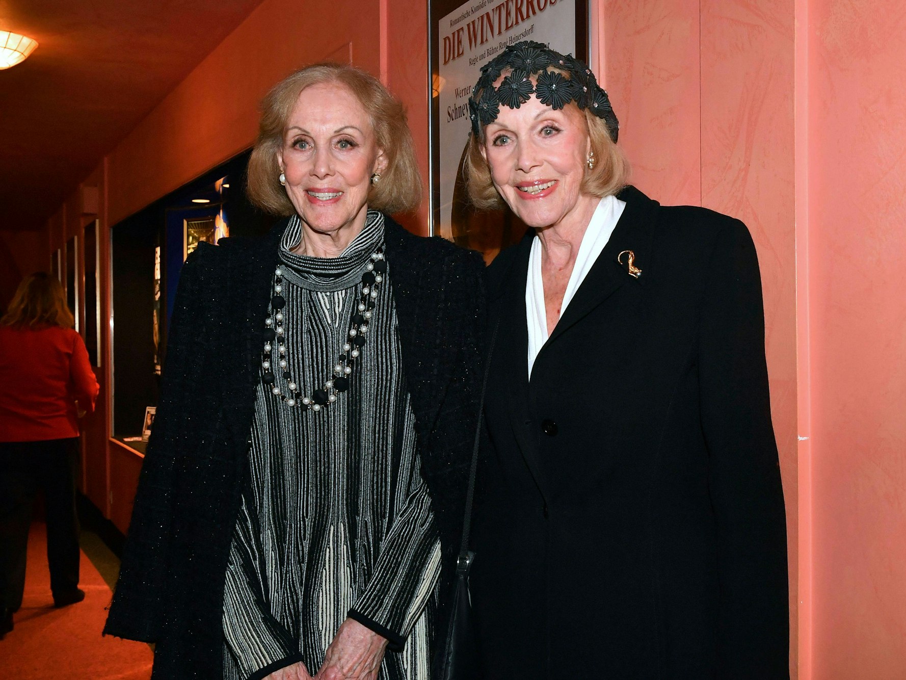 Zwei lächelnde ältere Frauen stehen nebeneinander vor einem Plakat in einem Innenraum