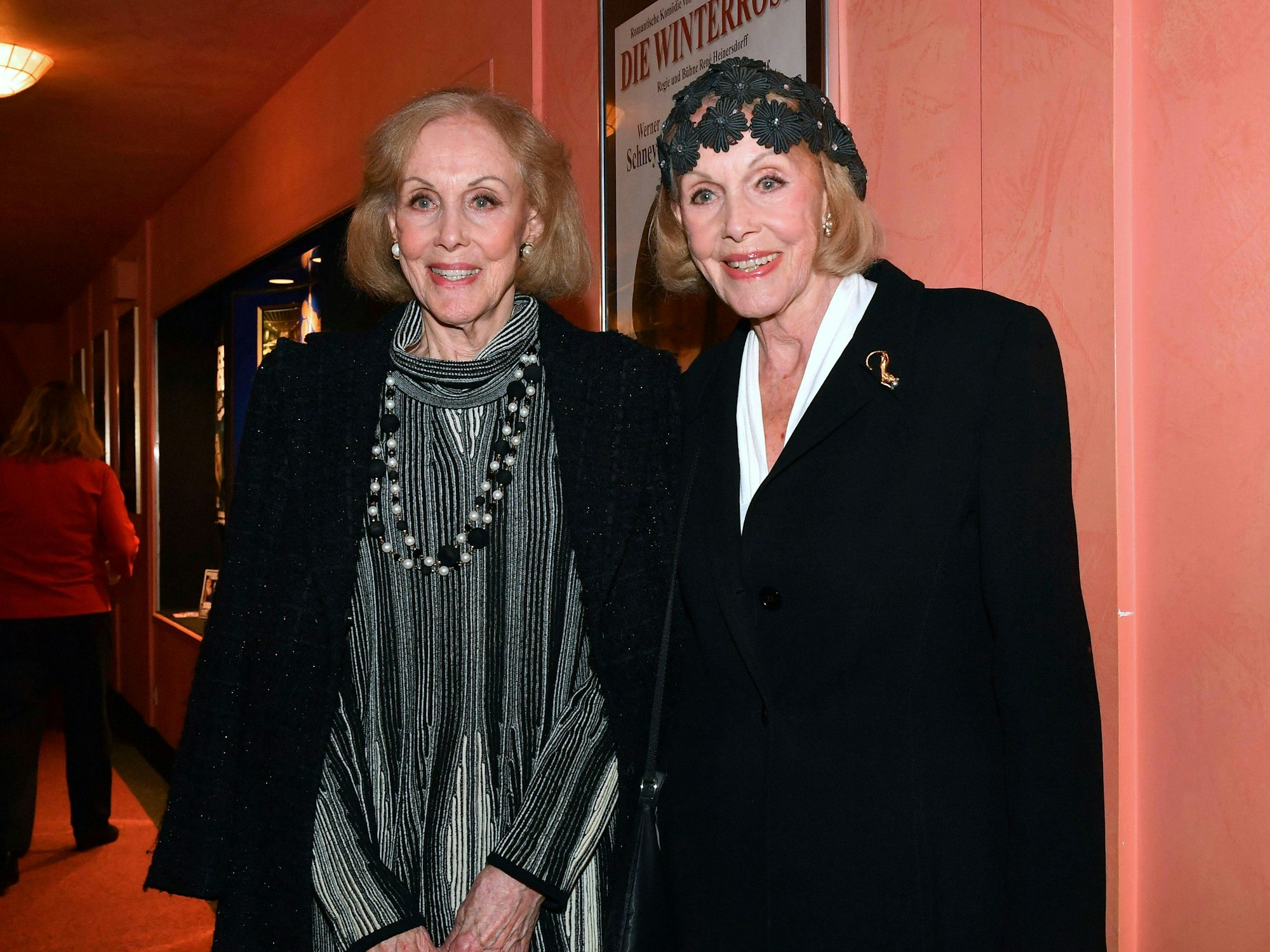 Zwei lächelnde ältere Frauen stehen nebeneinander vor einem Plakat in einem Innenraum