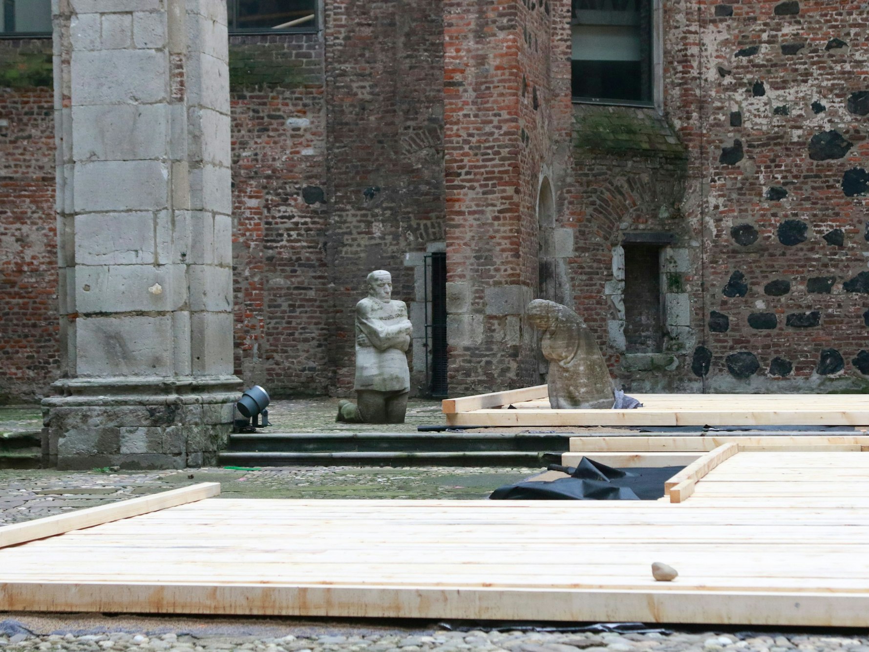 Innerhalb eines Gemäuers kniet eine Steinfigur vor einer zweiten, die beide Arme vor dem Oberkörper verschränkt hat.