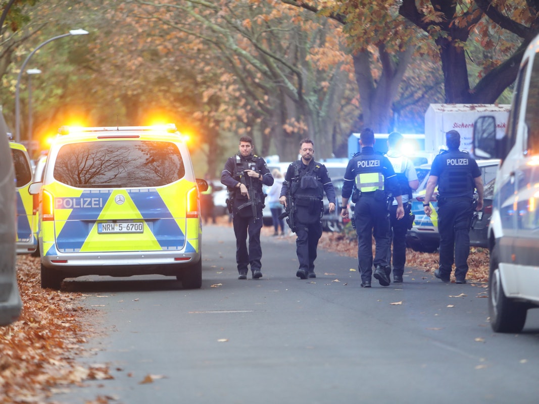 Einsatzkräfte laufen über eine Straße an einem Polizeifahrzeug vorbei.