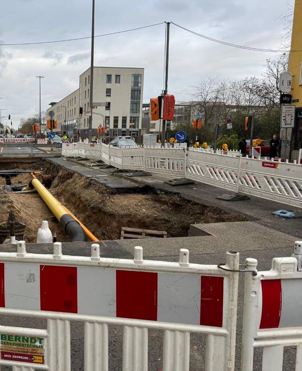 Auf der Fahrbahn einer Straße ist ein Graben mit Rohren, drumherum befindet sich ein Bauzaun.