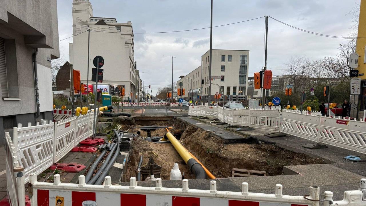 Auf der Fahrbahn einer Straße ist ein Graben mit Rohren, drumherum befindet sich ein Bauzaun.