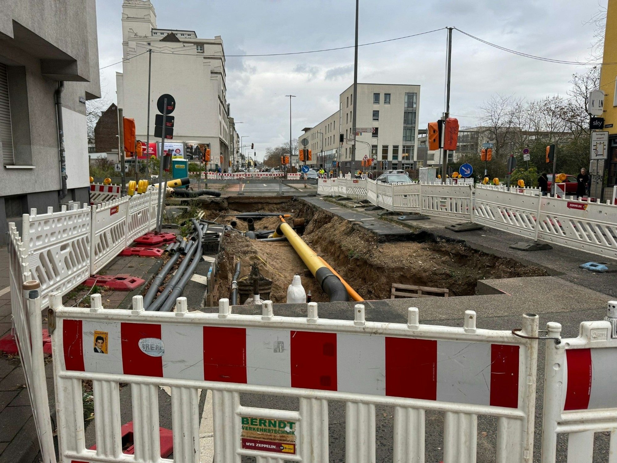 Auf der Fahrbahn einer Straße ist ein Graben mit Rohren, drumherum befindet sich ein Bauzaun.