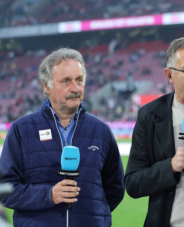 Achim Beierlorzer spricht bei Sport1 mit der Moderatorin und hält ein blaues Mikrofon dabei in der Hand. Neben ihm steht Peter Neururer.