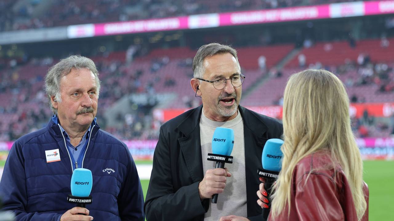 Achim Beierlorzer spricht bei Sport1 mit der Moderatorin und hält ein blaues Mikrofon dabei in der Hand. Neben ihm steht Peter Neururer.