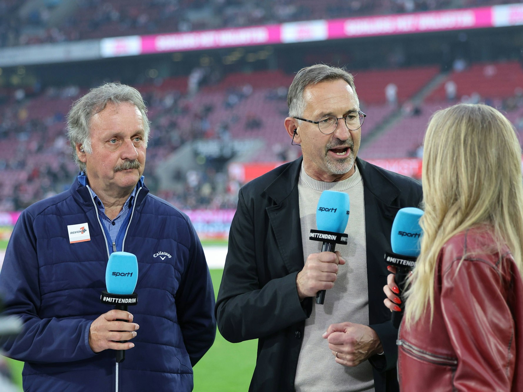 Achim Beierlorzer spricht bei Sport1 mit der Moderatorin und hält ein blaues Mikrofon dabei in der Hand. Neben ihm steht Peter Neururer.