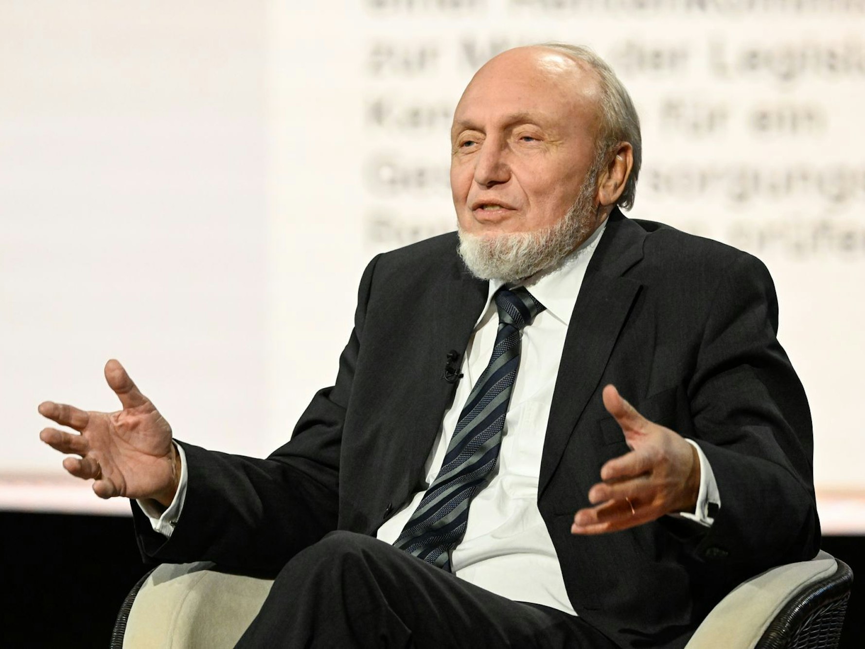 Ein Mann mit grauen Haaren und grauem Bart sitzt in einem Sessel und gestikuliert.