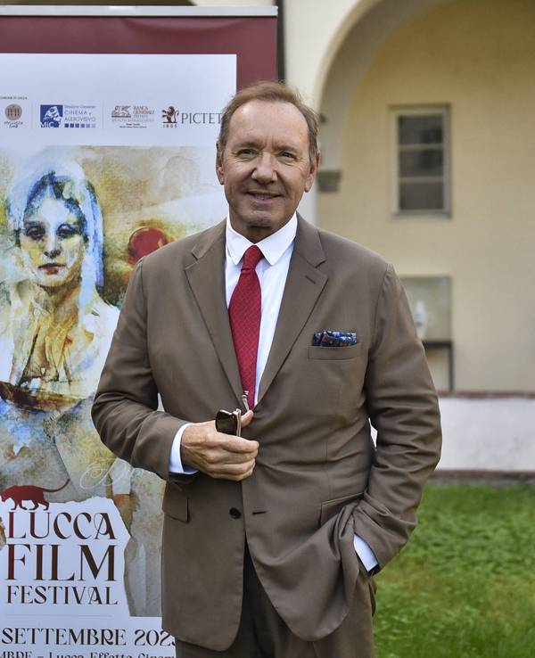Kevin Spacey lächelt und steht in einem braunen Anzug vor einem Banner des Lucca Film Festivals