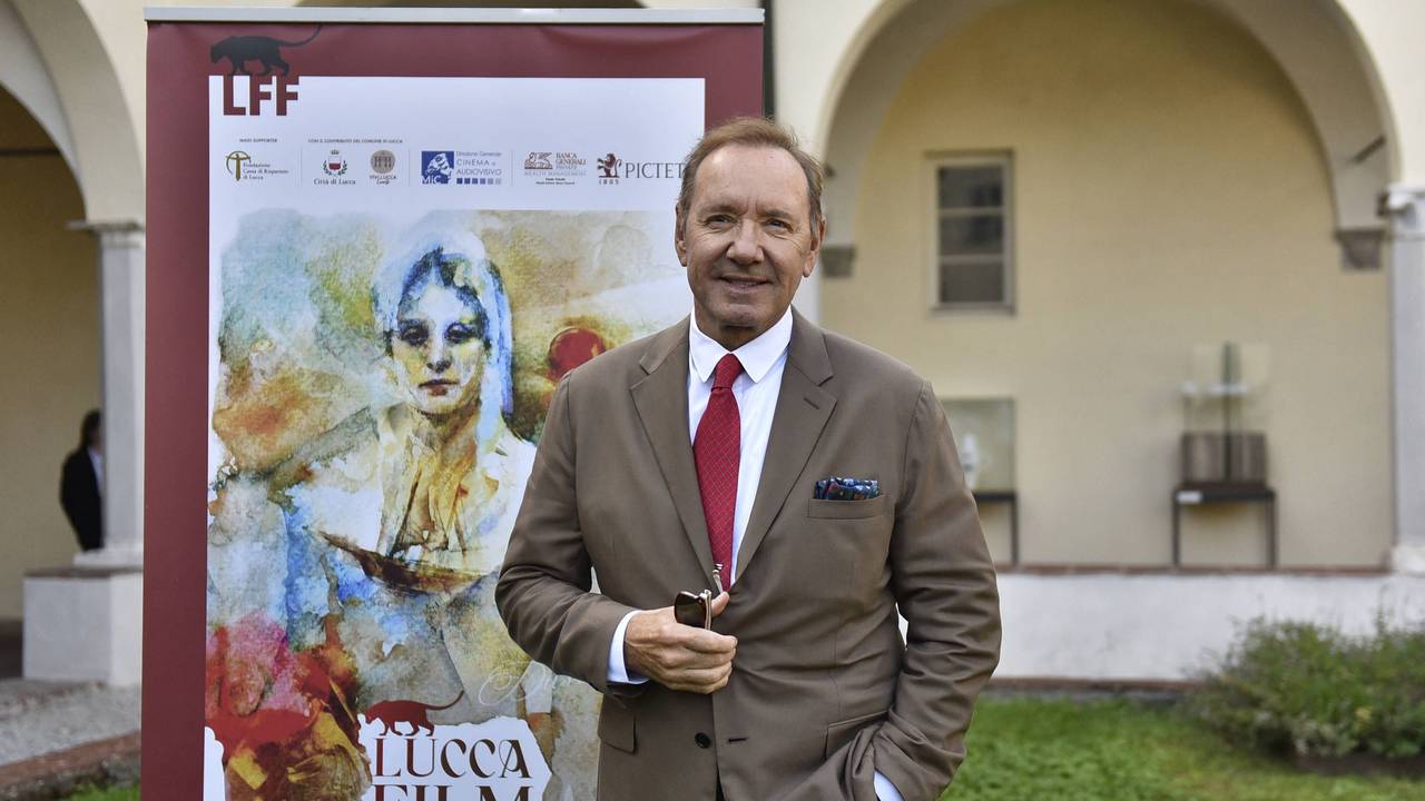 Kevin Spacey lächelt und steht in einem braunen Anzug vor einem Banner des Lucca Film Festivals