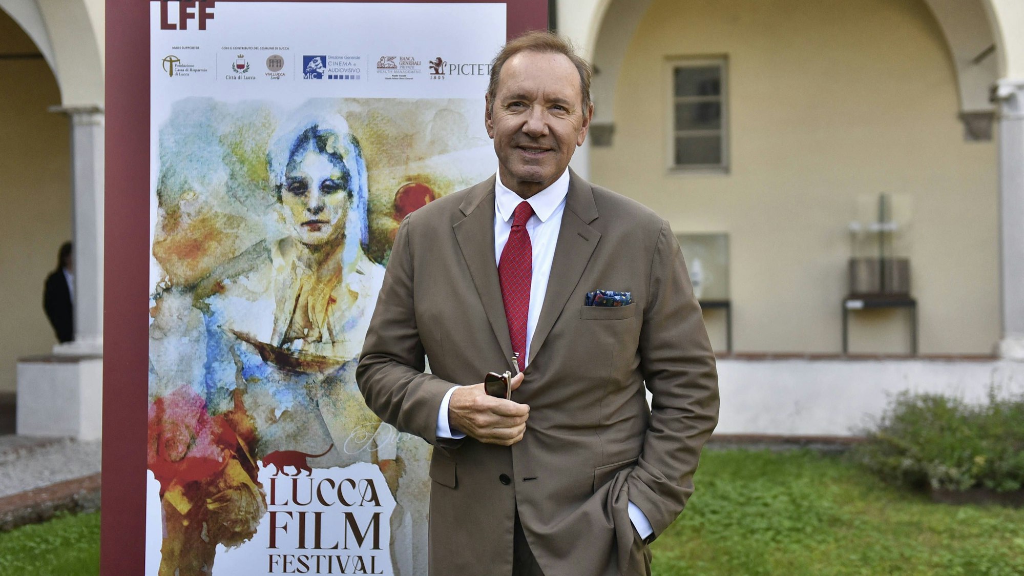 Kevin Spacey lächelt und steht in einem braunen Anzug vor einem Banner des Lucca Film Festivals