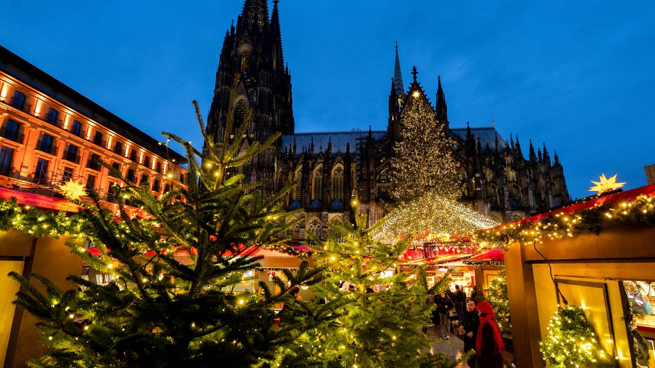 Vor dem Kölner Dom stehen beleuchtete Weihnachtsmarkt-Buden und Tannenbäume.