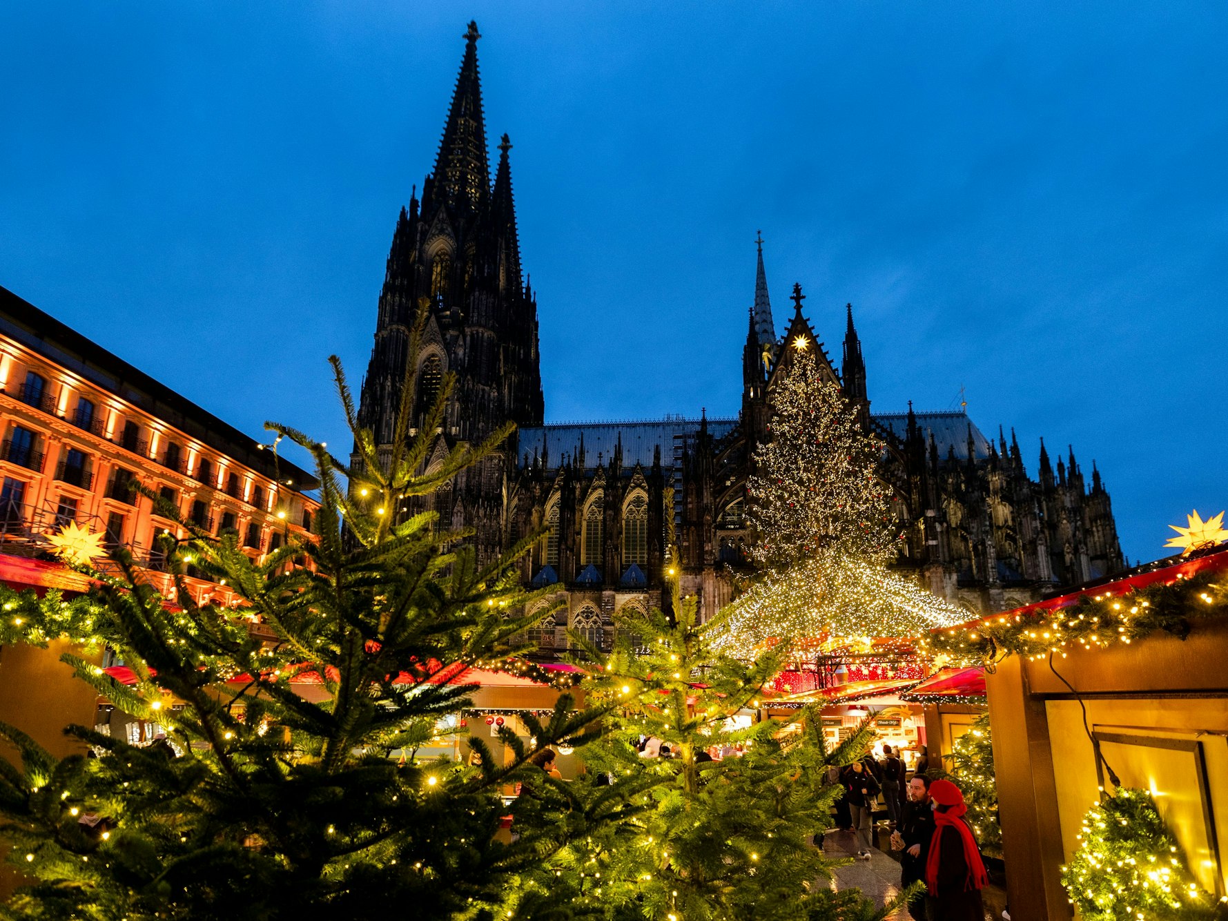 Vor dem Kölner Dom stehen beleuchtete Weihnachtsmarkt-Buden und Tannenbäume.