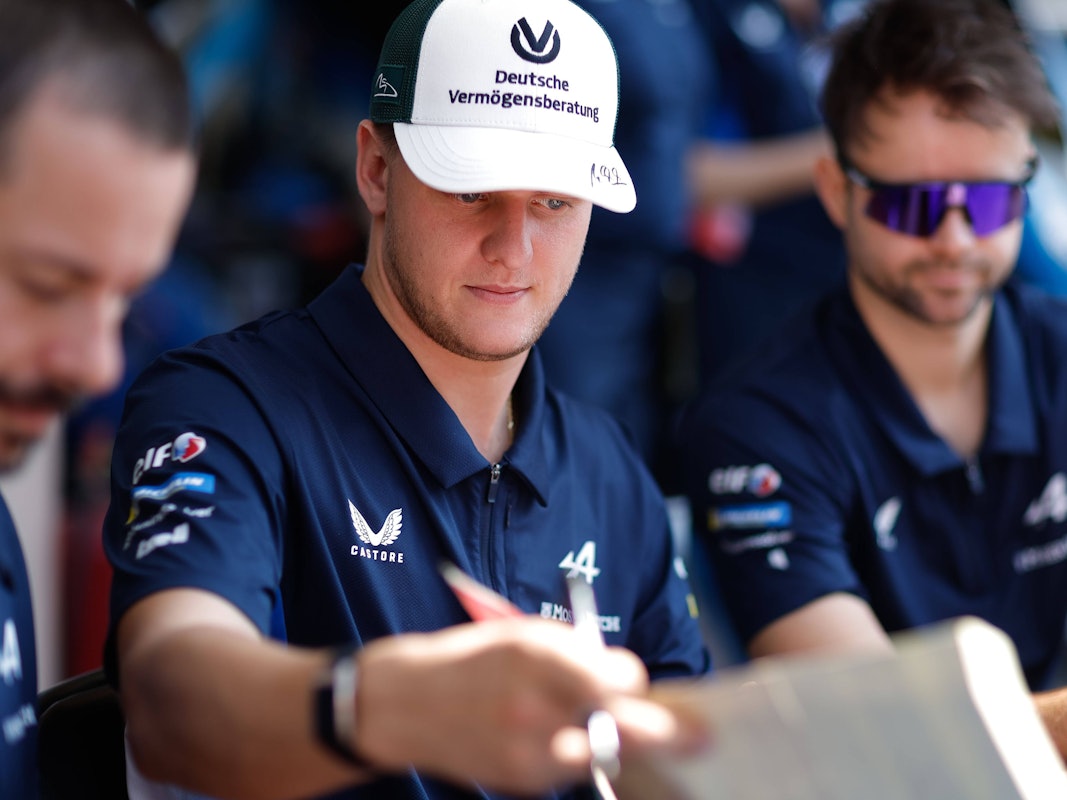 Mick Schumacher in Kappe und Polohemd unterschreibt etwas, umgeben von zwei Männern