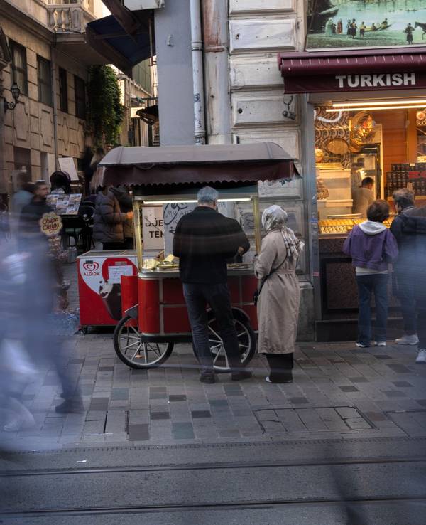 Eine Straßenszene, ein Paar steht vor einem mobilen Essensstand, daneben ein Geschäft für türkische Spezialitäten, unkenntlich gemachte Menschen laufen vorbei.