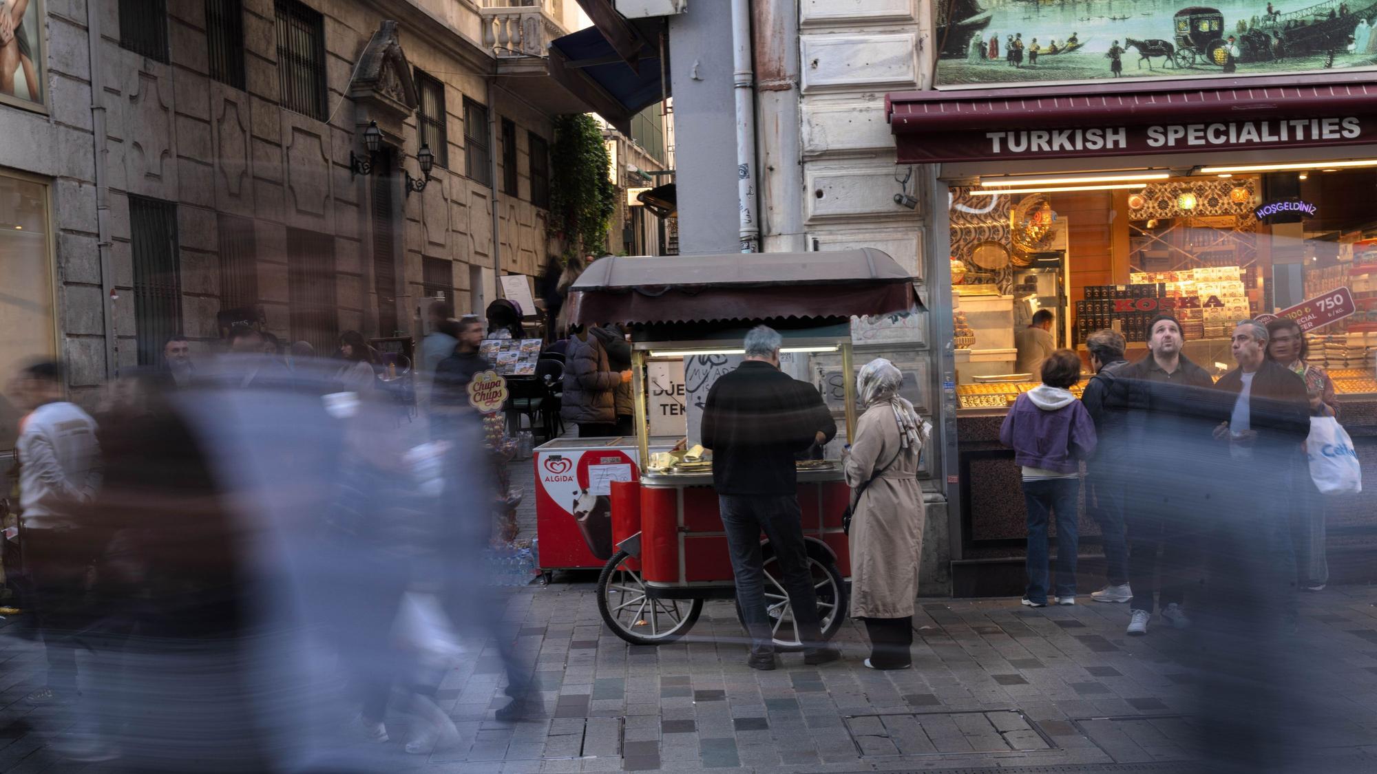 Eine Straßenszene, ein Paar steht vor einem mobilen Essensstand, daneben ein Geschäft für türkische Spezialitäten, unkenntlich gemachte Menschen laufen vorbei.