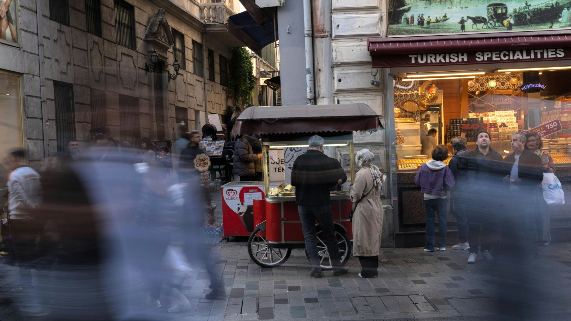 Eine Straßenszene, ein Paar steht vor einem mobilen Essensstand, daneben ein Geschäft für türkische Spezialitäten, unkenntlich gemachte Menschen laufen vorbei.