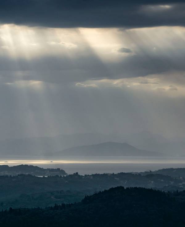Blick auf Festland und Inseln, der Himmel ist dunkel, zwischen den Wolken kommen einzelne Sonnenstrahlen durch.