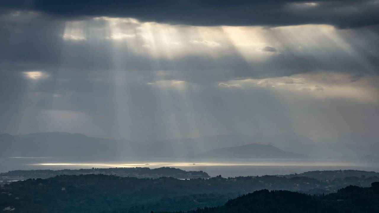 Blick auf Festland und Inseln, der Himmel ist dunkel, zwischen den Wolken kommen einzelne Sonnenstrahlen durch.