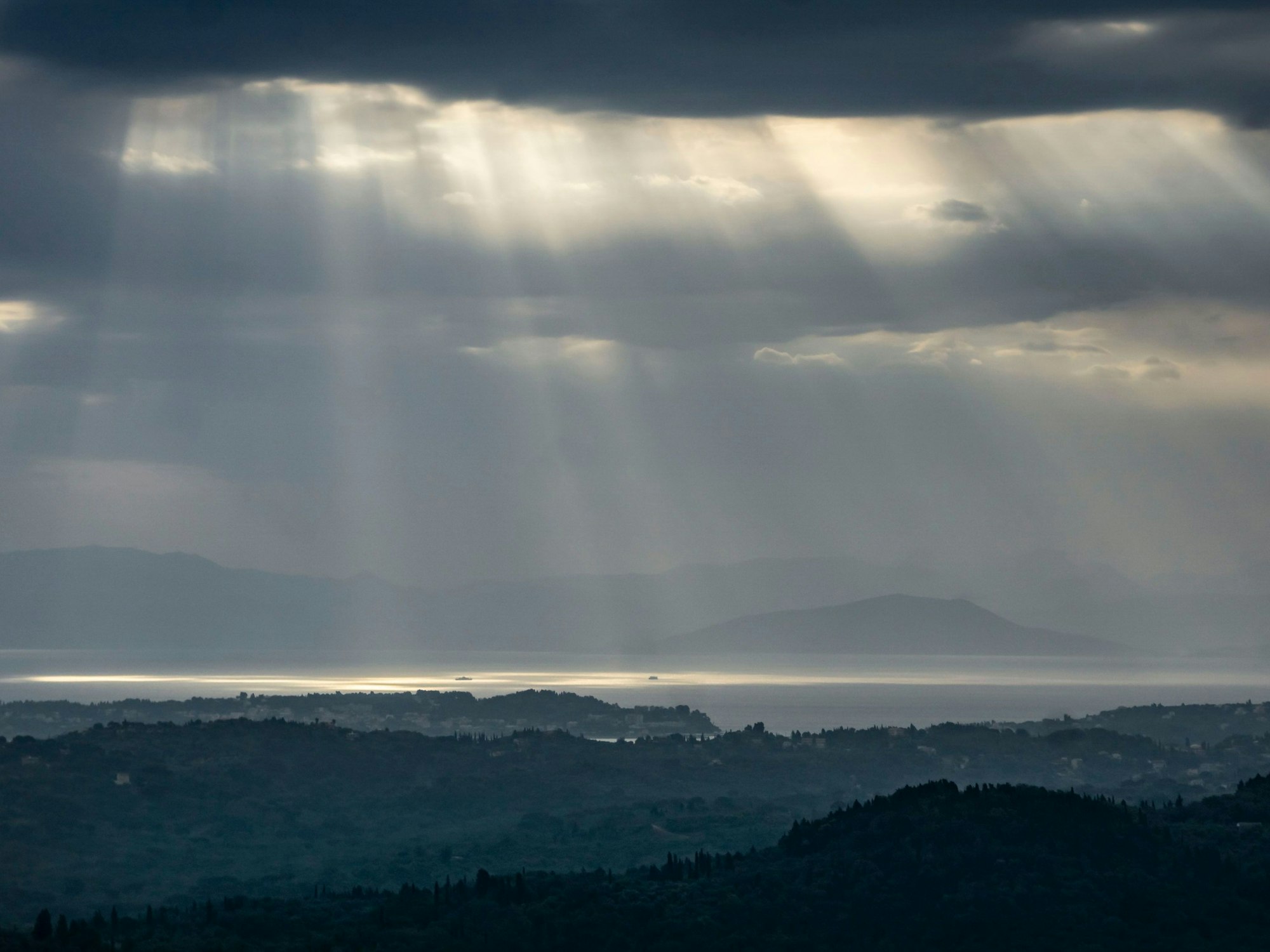 Blick auf Festland und Inseln, der Himmel ist dunkel, zwischen den Wolken kommen einzelne Sonnenstrahlen durch.