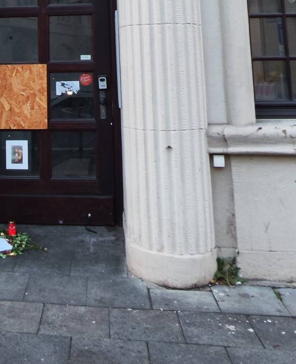 Vor einer Tür zwischen zwei Säulen liegen Blumen vor einem Bilderrahmen und einer Kerze.