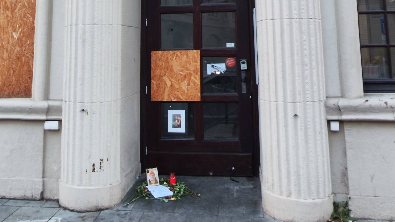 Vor einer Tür zwischen zwei Säulen liegen Blumen vor einem Bilderrahmen und einer Kerze.