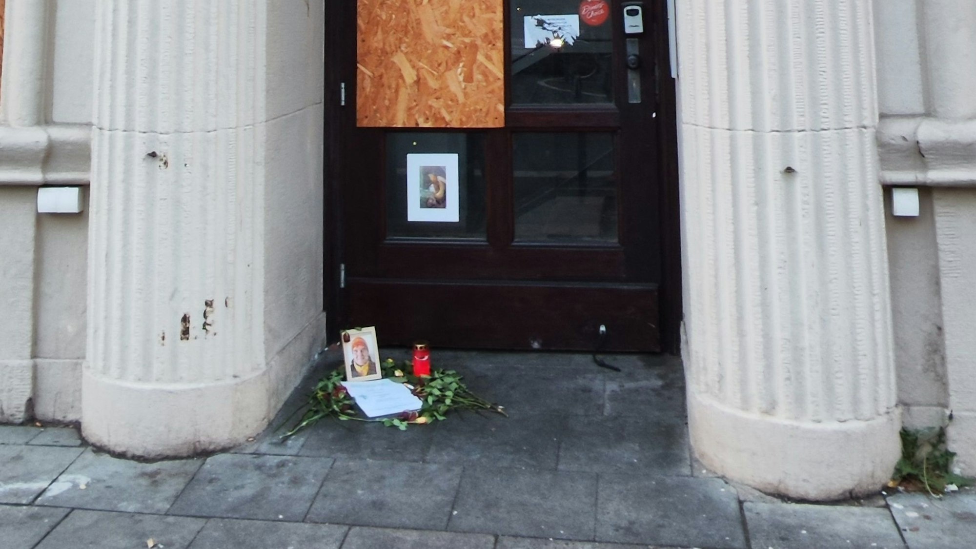 Vor einer Tür zwischen zwei Säulen liegen Blumen vor einem Bilderrahmen und einer Kerze.