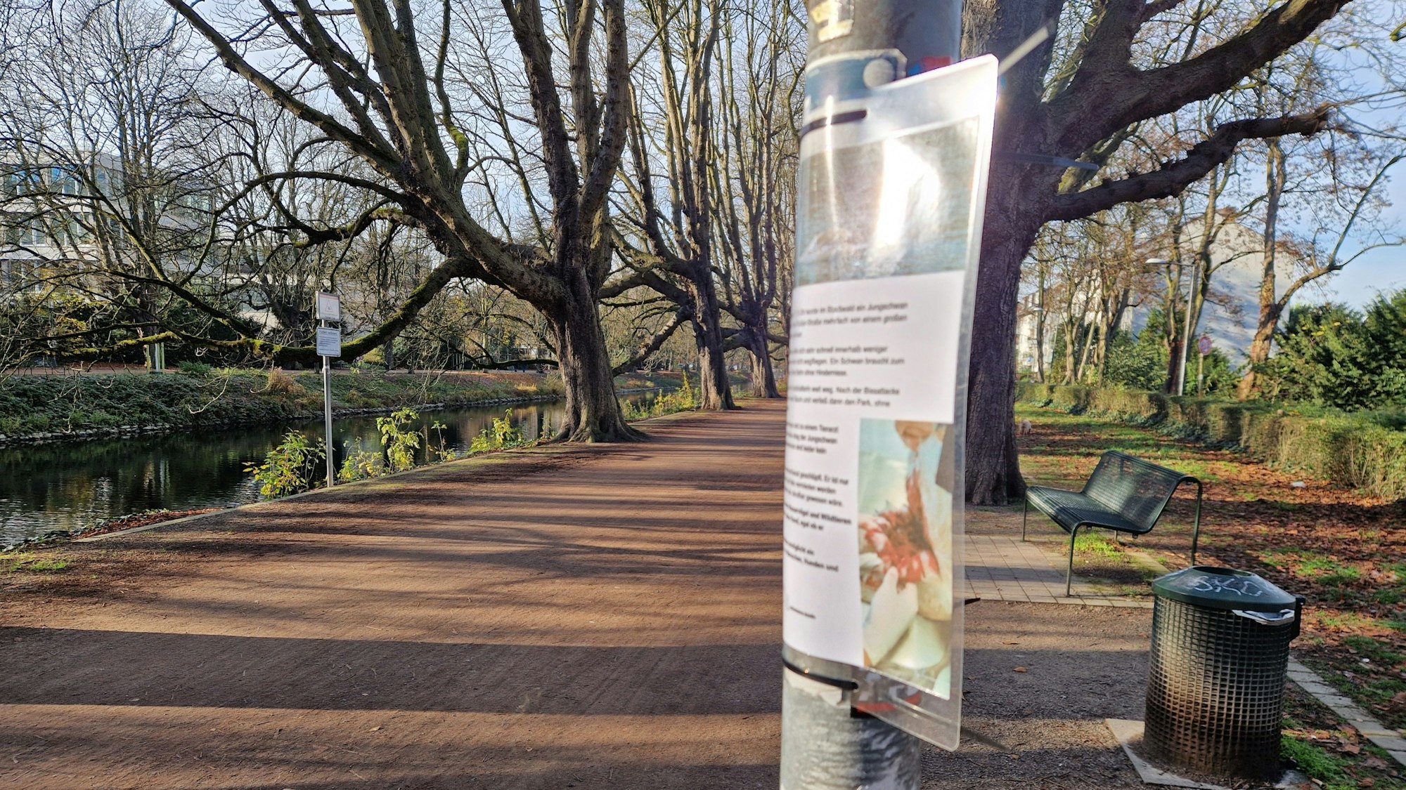 Am Clarenbachkanal in Köln-Lindenthal hängt ein laminiertes Schild an einem Metallpfal.
