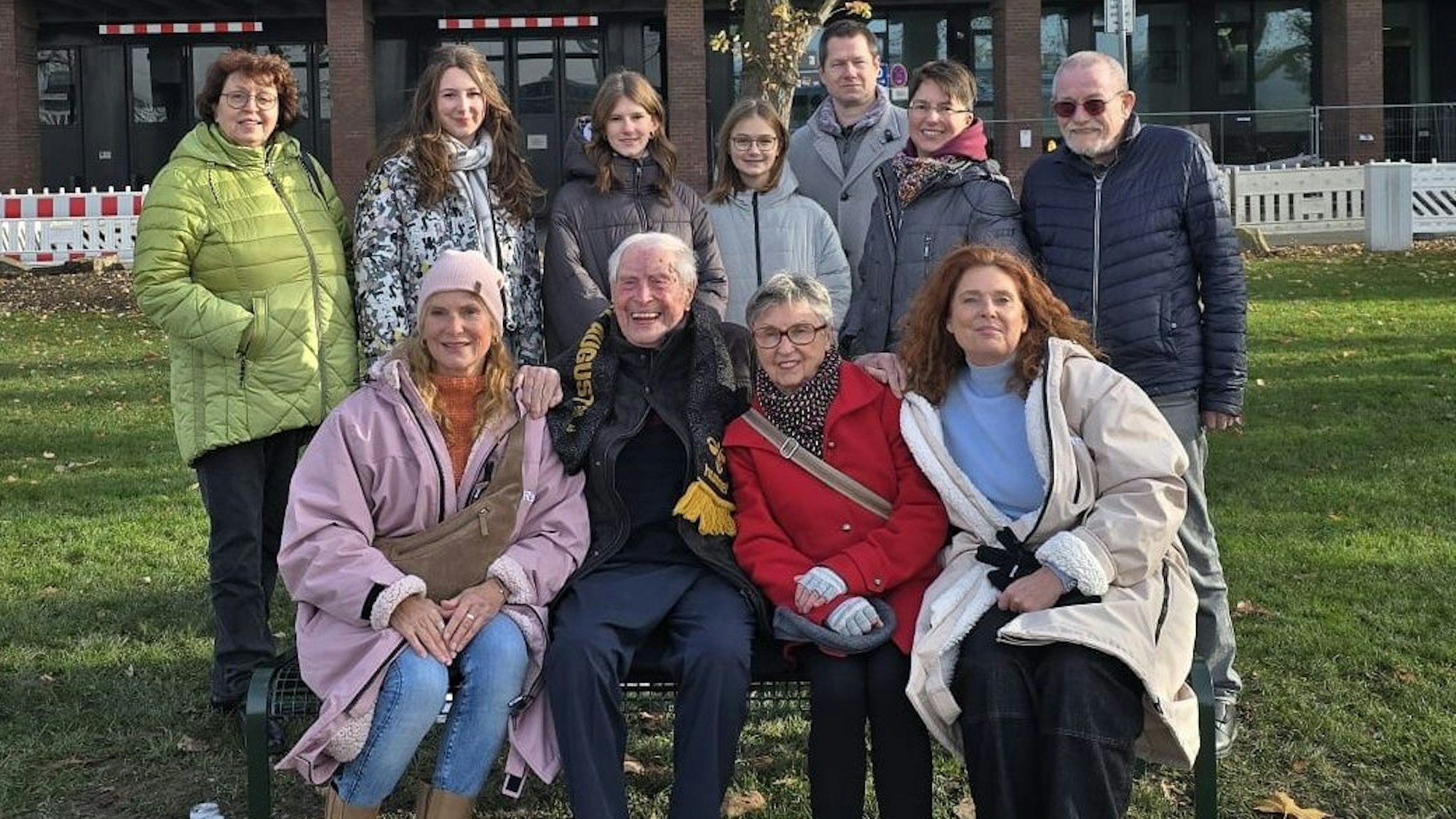 Ludwig Sebus sitzt umrahmt von seiner Familie auf seiner Bank am Rheinufer.