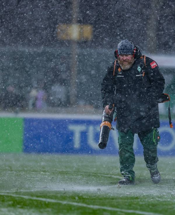 Ein Helfer mit Laubbläser pustet Schnee beim Spiel der Frauen-Bundesliga vom Feld.