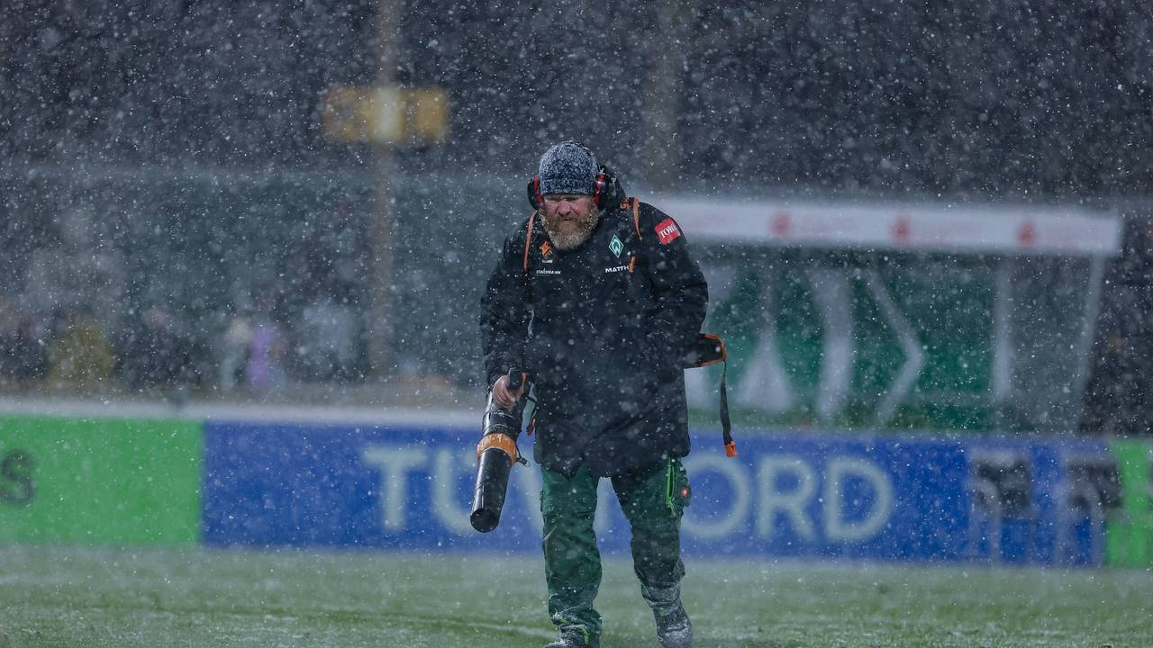 Ein Helfer mit Laubbläser pustet Schnee beim Spiel der Frauen-Bundesliga vom Feld.