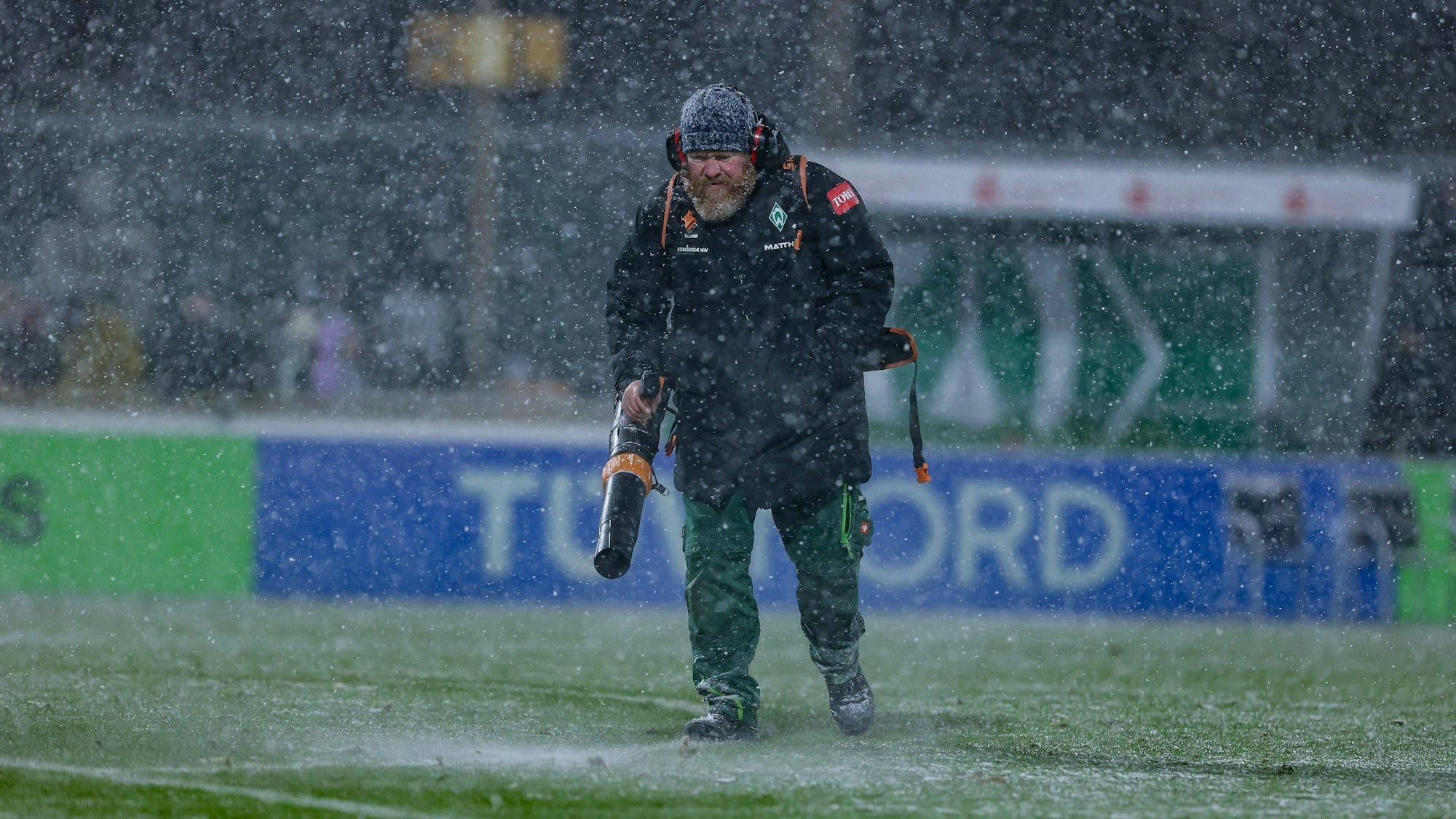 Ein Helfer mit Laubbläser pustet Schnee beim Spiel der Frauen-Bundesliga vom Feld.