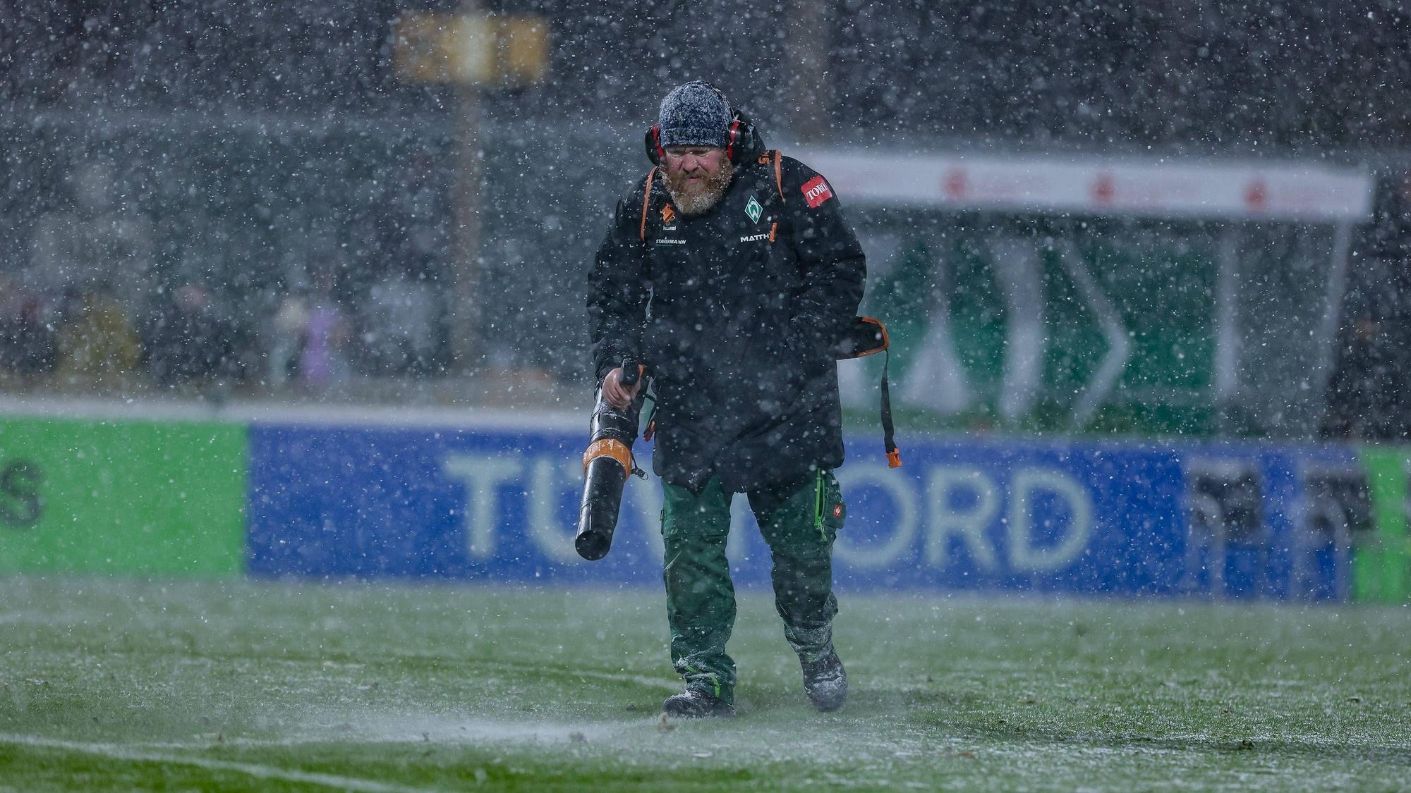 Ein Helfer mit Laubbläser pustet Schnee beim Spiel der Frauen-Bundesliga vom Feld.