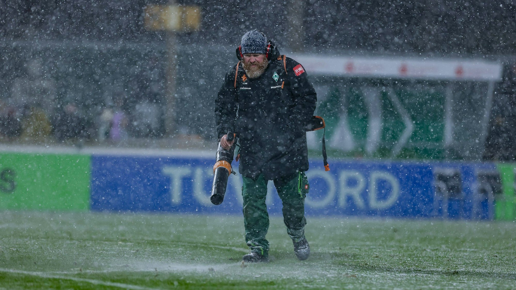 Ein Helfer mit Laubbläser pustet Schnee beim Spiel der Frauen-Bundesliga vom Feld.