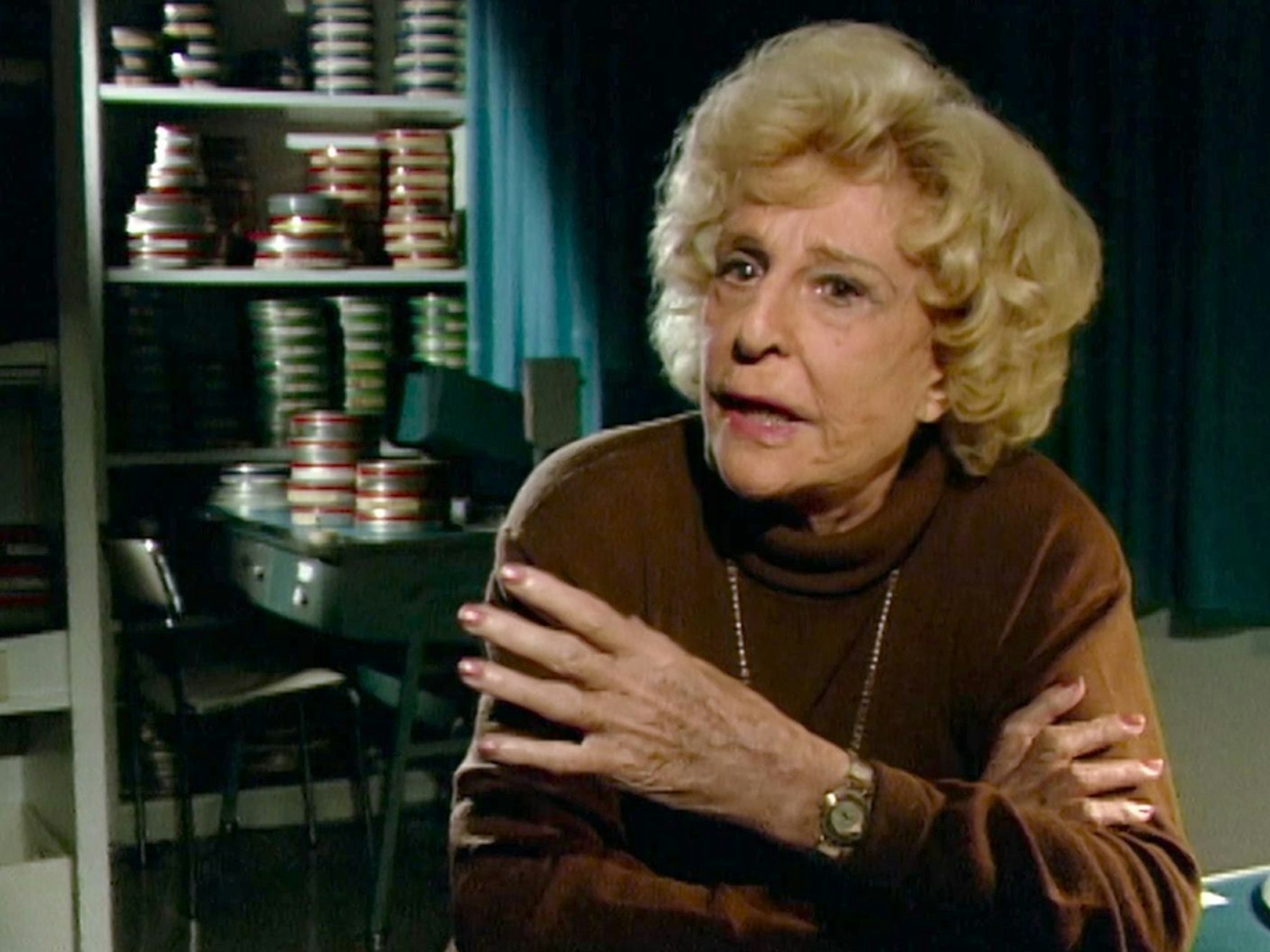 Eine ältere Frau sitzt vor einem Regal, in dem sich Filmrollen stapeln.