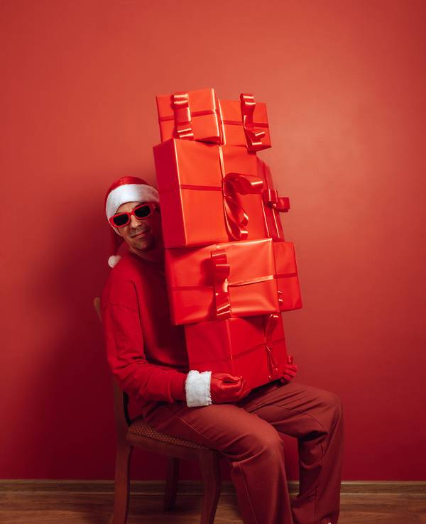 Nikolaus in rotem Outfit und Sonnenbrille, mit großem Stapel Geschenke vor rotem Hintergrund