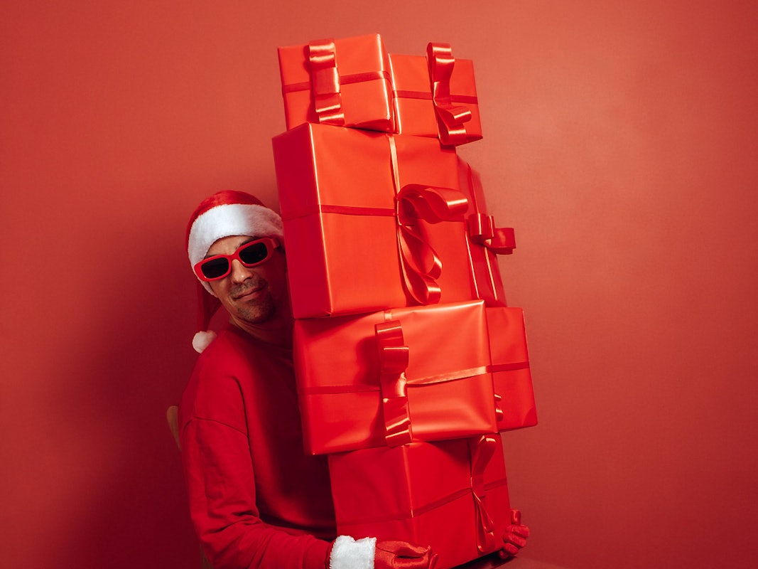 Nikolaus in rotem Outfit und Sonnenbrille, mit großem Stapel Geschenke vor rotem Hintergrund